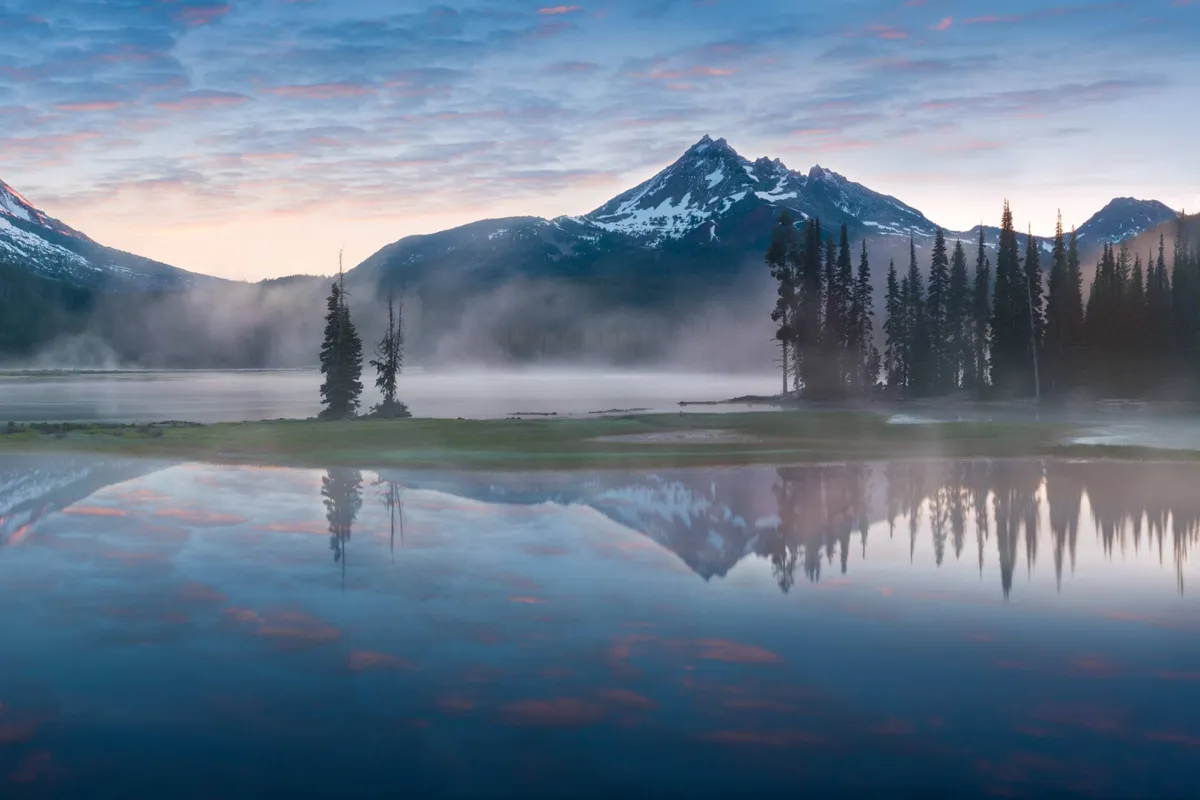 A tranquil mountain lake surrounded by tall evergreens, with mountains rising in the background and a soft sky overhead. The scene suggests early morning or evening, perfect for peaceful hikes, birdwatching, or simply relaxing by the water.