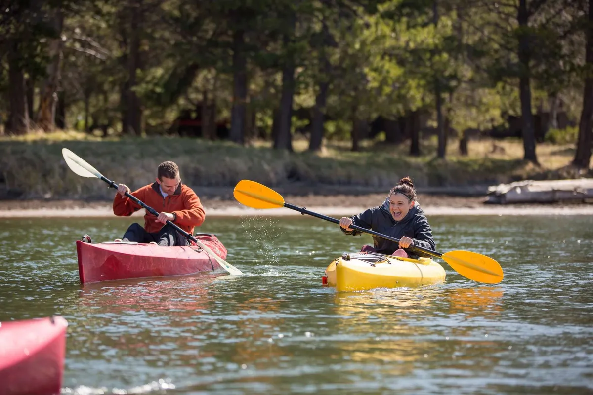 On a sparkling river, two kayakers paddle vibrant kayaks amid lush green trees. The scene is lively and inviting, highlighting the fun and active outdoor pursuits available in the Sunriver area.