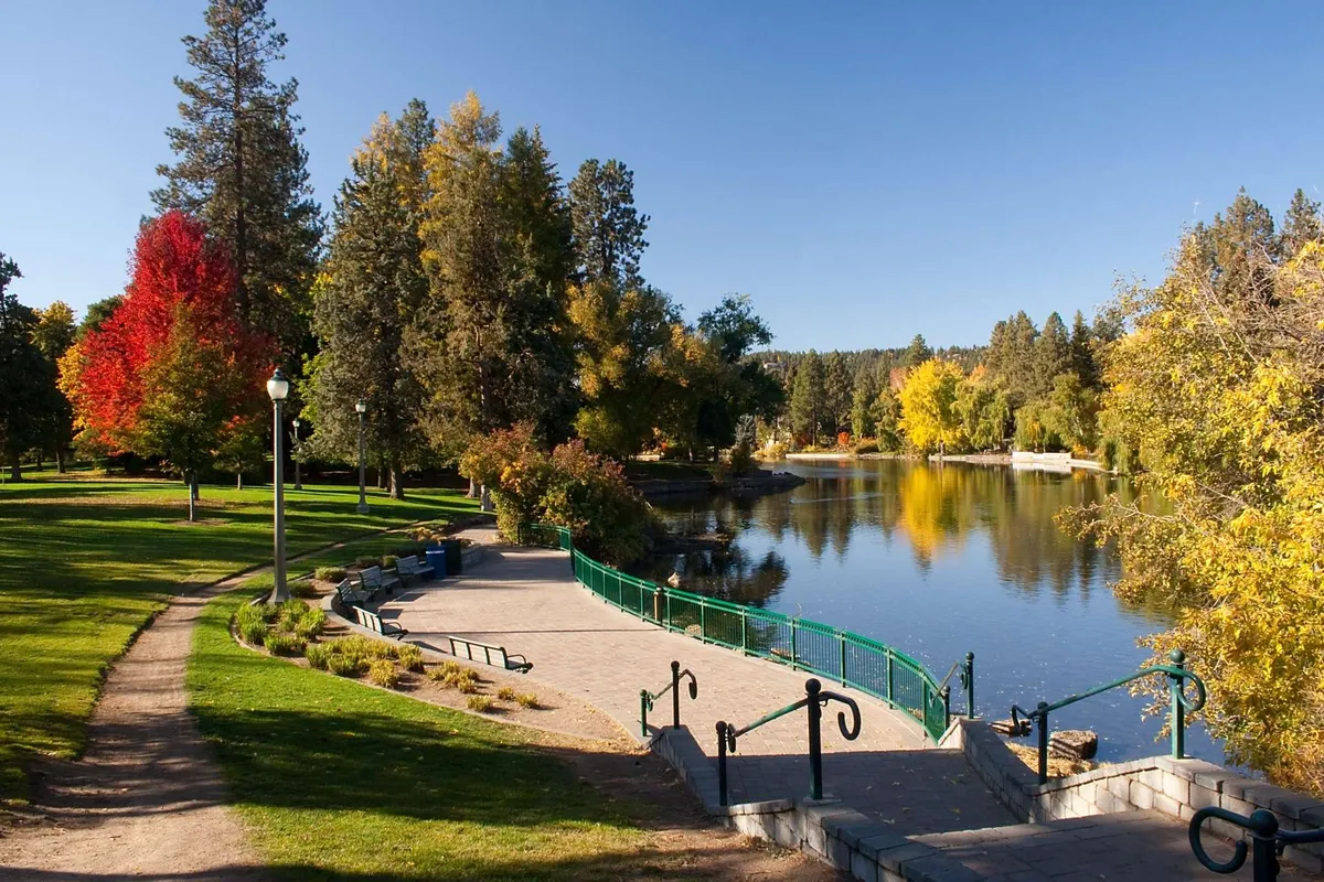A tranquil riverside scene reveals a winding walking path shaded by colorful trees and lush greenery. Park benches invite relaxation, and the calm waters offer a peaceful backdrop, ideal for family strolls or moments of reflection in nature.