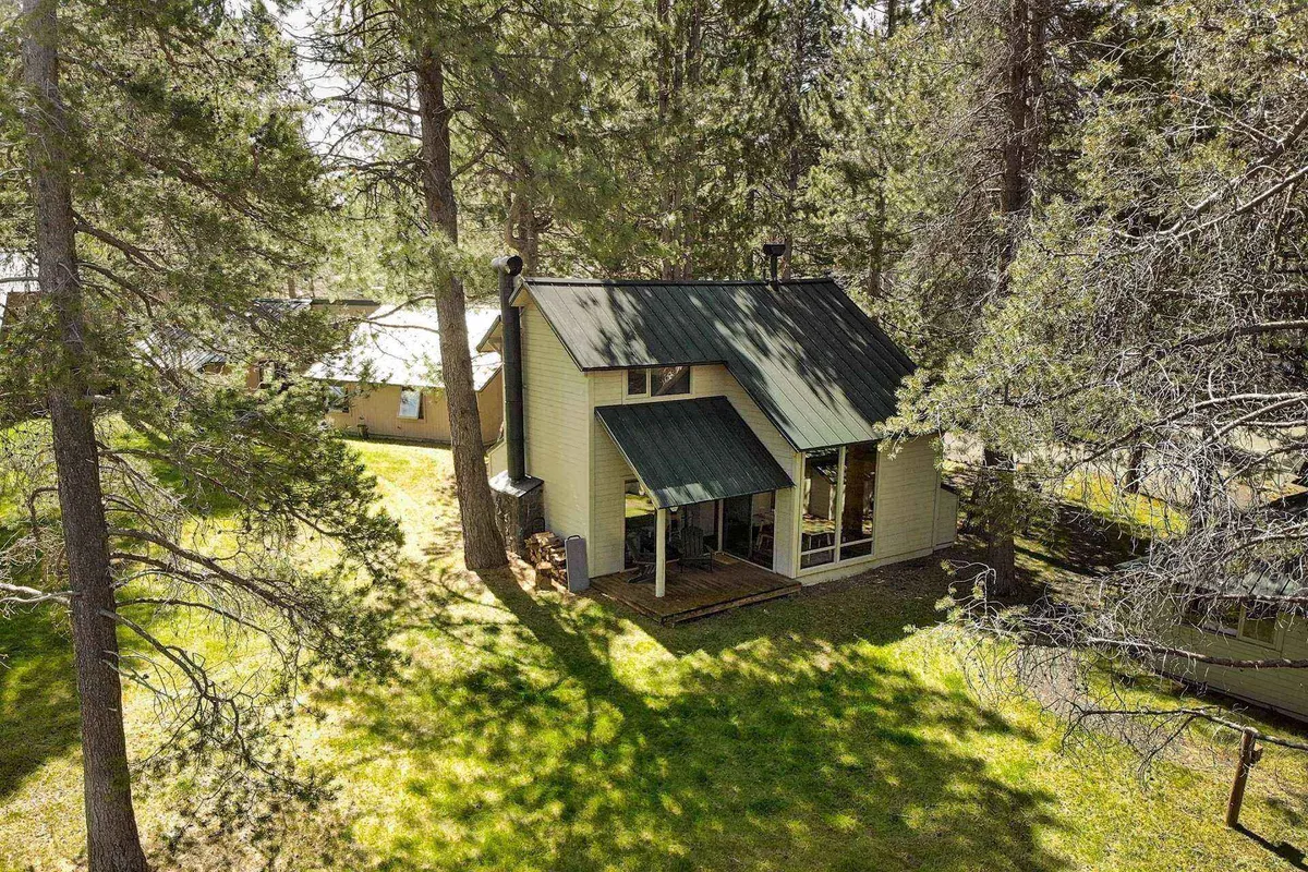 A charming wooden house surrounded by mature trees and a green lawn, with a welcoming front porch that hints at outdoor relaxation and access to nature. The scene is tranquil, inviting guests to imagine stepping outside to nearby trails.
