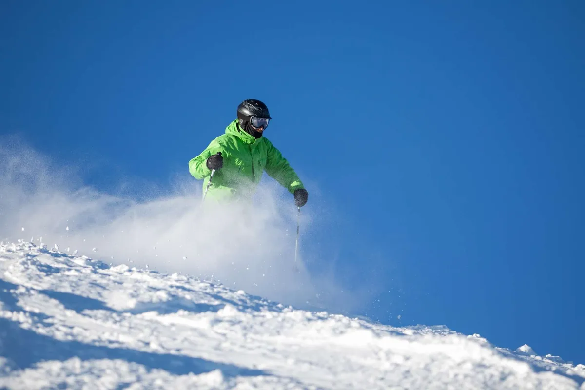 An action-filled shot of a skier making a thrilling descent down a snow-covered slope, framed by a brilliant blue sky. The skier is captured mid-motion, emphasizing energetic outdoor pursuits.