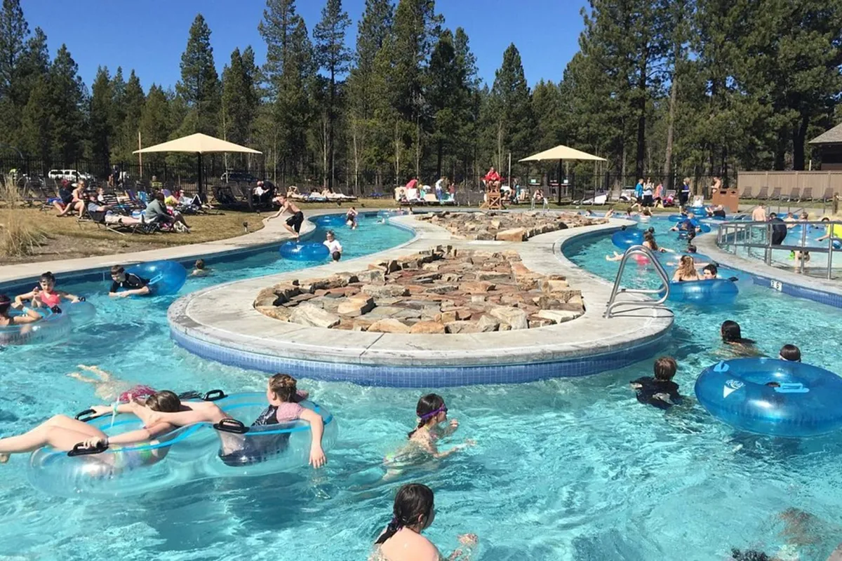 A sunlit swimming pool with a natural rock formation, poolside loungers shaded by umbrellas, and people floating on tubes. The mood is relaxed yet joyful, suggesting lazy afternoons and togetherness by the water.