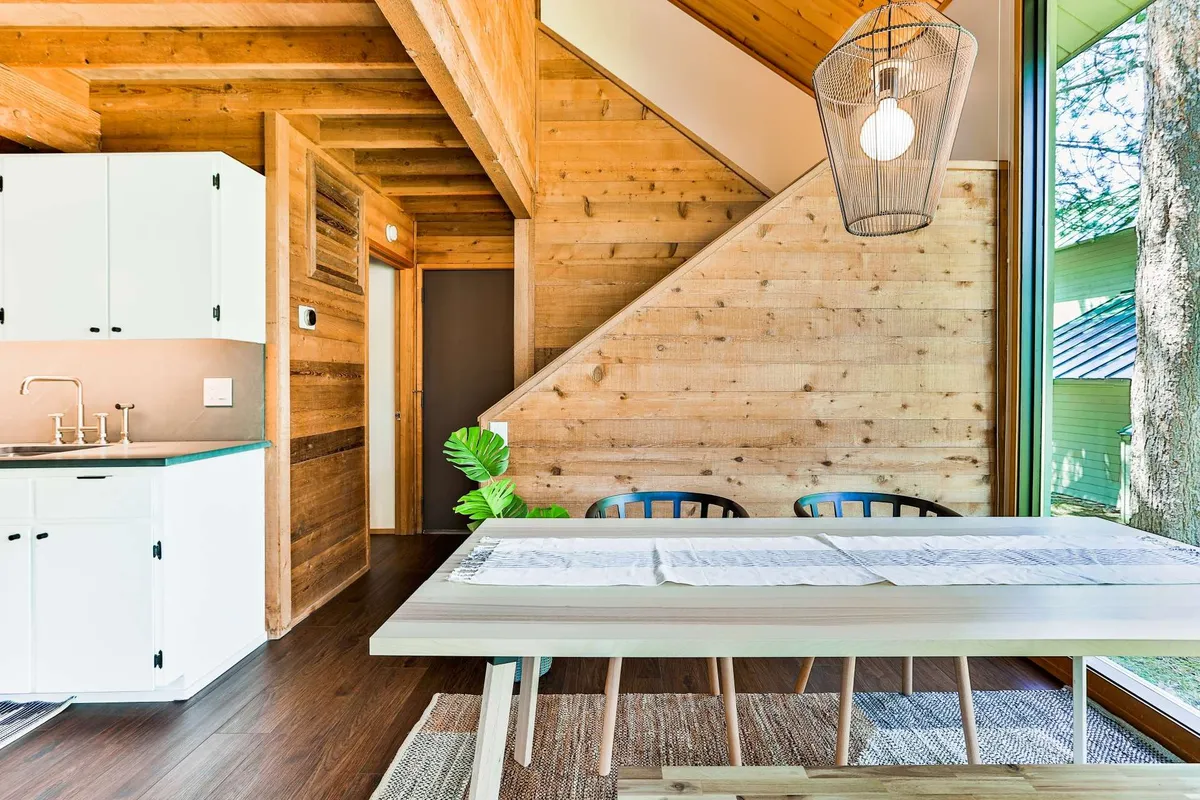 A spacious, sun-drenched dining area anchored by a wooden dining table next to the kitchen and large windows. The area features a staircase leading upward, modern kitchen appliances, and plenty of space for group meals or family activities.