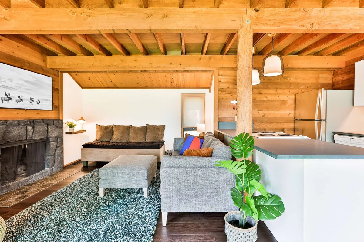 A warm, inviting living area featuring a wood-burning fireplace as its centerpiece. Plush sofas are situated nearby, with a kitchen island visible in the background. Large windows allow natural light to fill the space, and tasteful artwork and a houseplant add touches of comfort and style.
