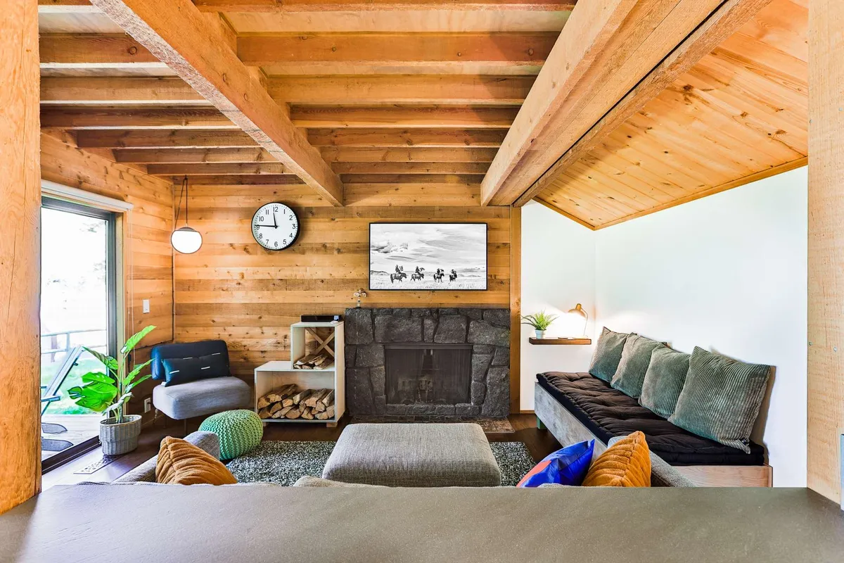 The hero image showcases the cabin's inviting living room, centered around a striking stone fireplace beneath rustic wooden beams. Natural light pours in through expansive windows, offering a view of the outdoors, while plush seating and tasteful artwork complete the warm, welcoming atmosphere.