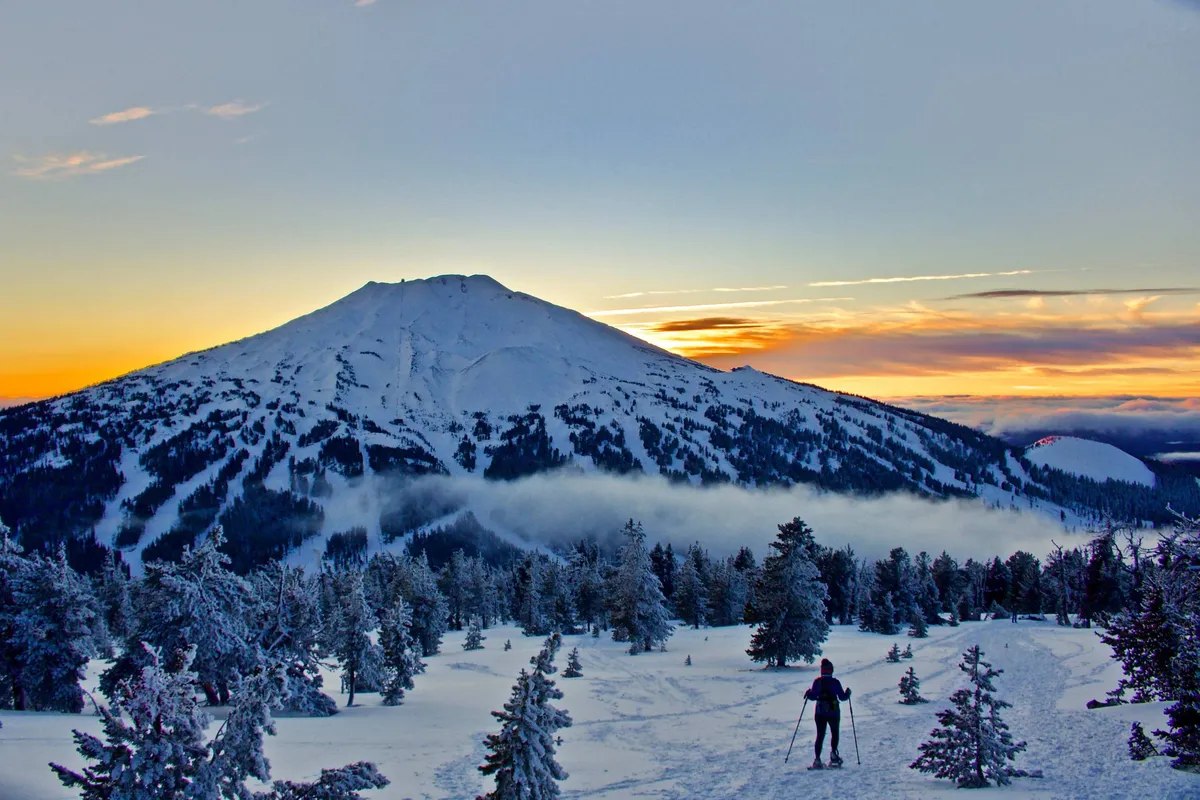 A sweeping view of a snow-covered mountain beneath a sunset sky, with a skier in the foreground poised on pristine powder. The scene captures the serenity and grandeur of winter adventure, blending natural beauty with action.